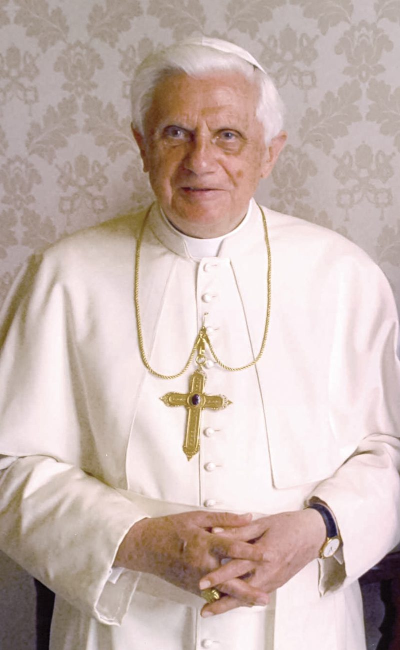 Pope Benedict XVI in white cassock, smiling, hands folded.