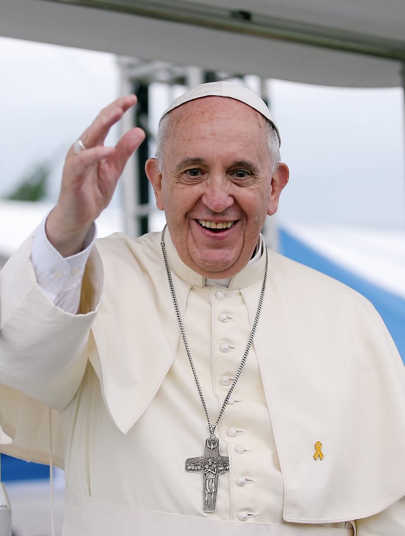 Pope Francis in white cassock, smiling and waving, outdoors.