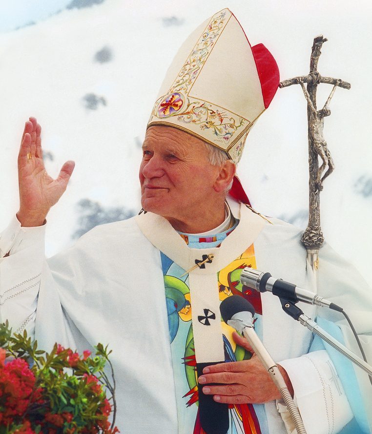 Pope St John Paul II in white vestments and mitre, arms raised in greeting at an outdoor Mass.