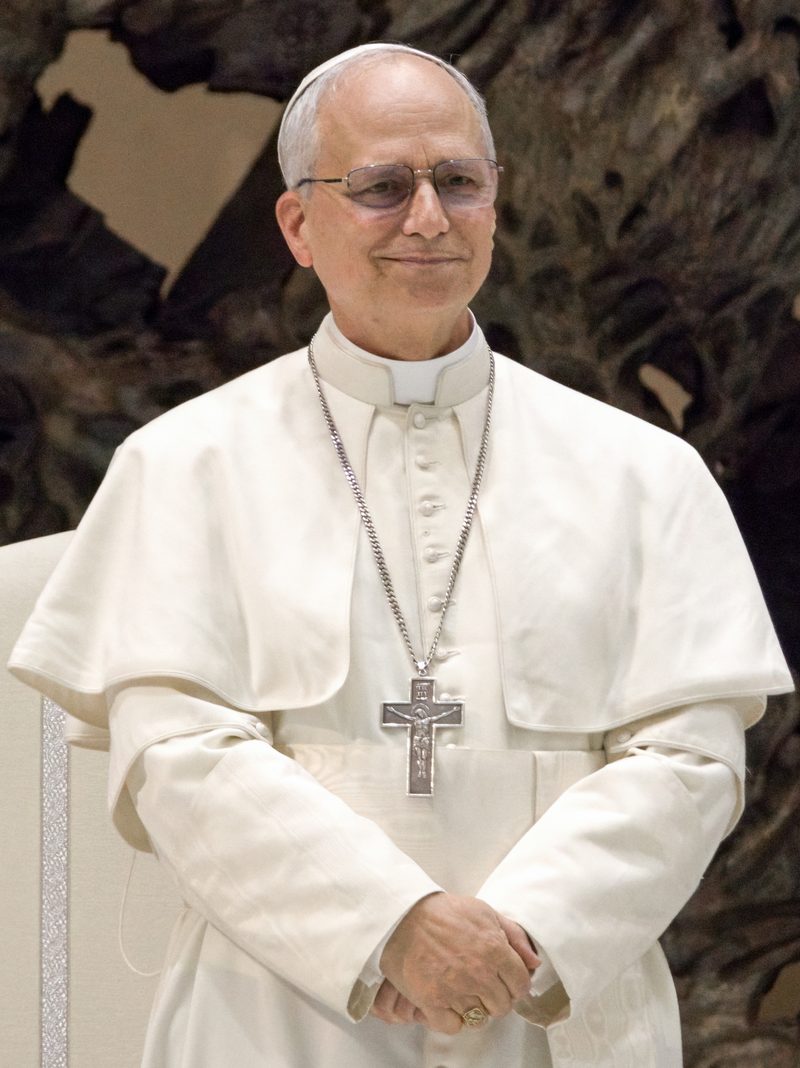 Pope Leo XIV in white, smiling, outdoors.