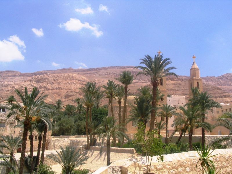 The Monastery of St Anthony in the Egyptian desert: palm trees and pale buildings at the foot of bare red mountains.