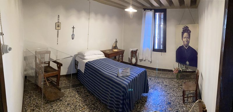 The simple room of St Josephine Bakhita at Schio: a narrow bed with a blue cover, a chair and a crucifix.