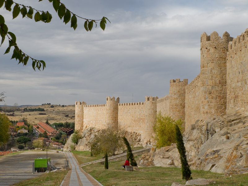 The medieval walls of Avila, Spain: golden stone ramparts with round towers under a blue sky.